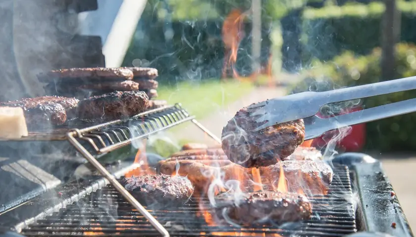 Des hamburgers grillent sur un barbecue à gaz. La fumée s'élève, les flammes dansent. Un hamburger est retourné avec une pince.