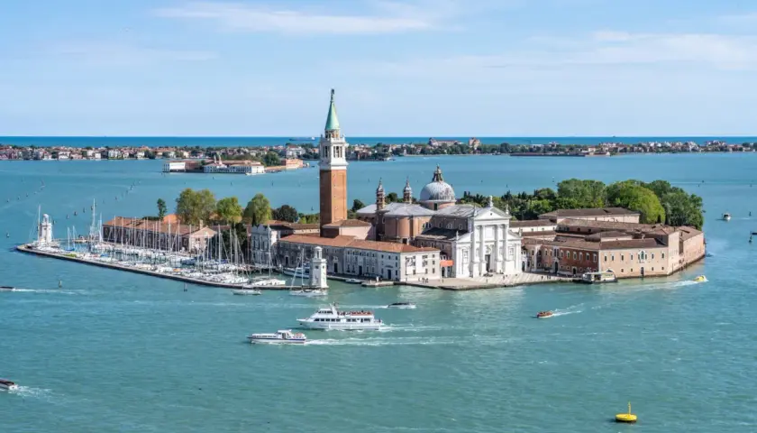 Widok na wyspę San Giorgio Maggiore w Wenecji, z kościołem i dzwonnicą. Wokół pływają łodzie, w tym prom.