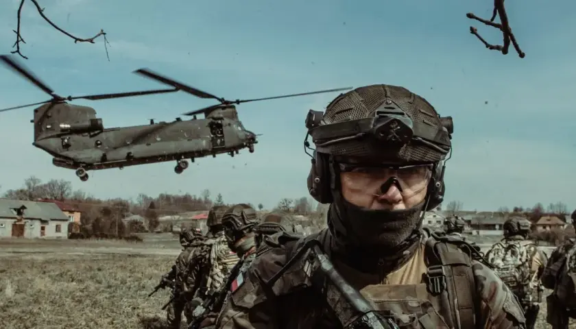 Żołnierze w pełnym rynsztunku, gotowi do akcji podczas szkolenia wojskowego. W tle widać helikopter transportowy.