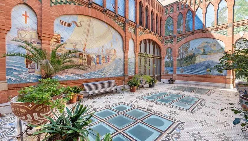 Patio de la Casa Navàs de Reus, con murales cerámicos que narran historias marítimas y paisajes, suelo de mosaico y plantas.