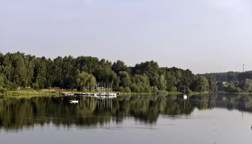 Spokojne jezioro Przeczyckie z odbijającymi się w wodzie drzewami i żaglówkami przy brzegu.
