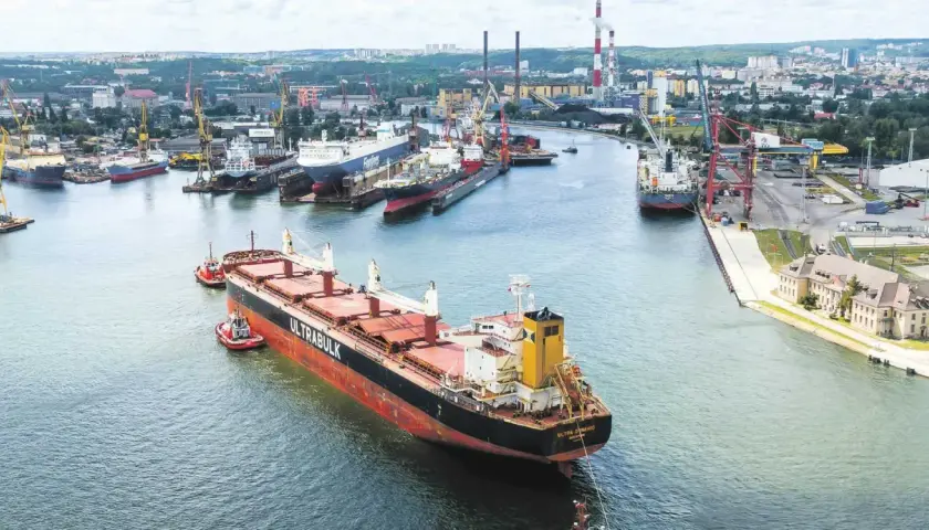 Duży statek towarowy "ULTRABULK" manewruje w porcie z pomocą holowników, w tle stocznie i miasto.