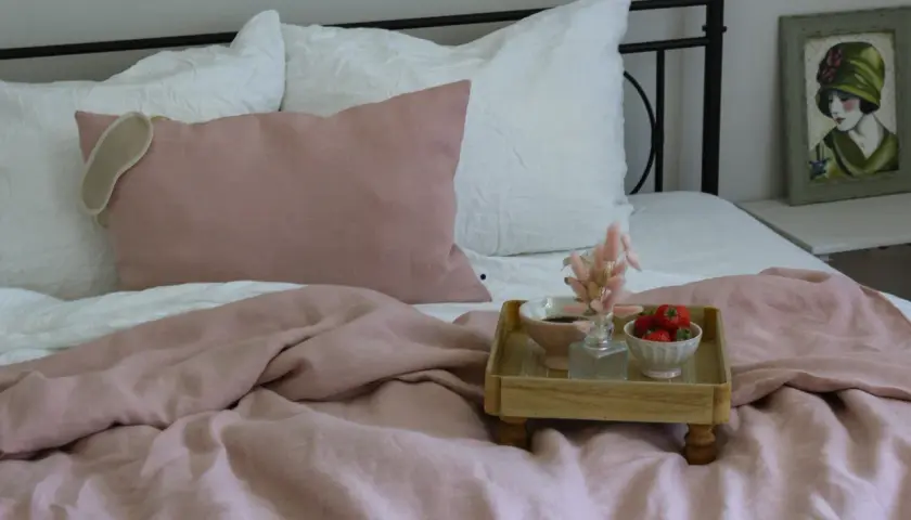 Ein rosafarbenes Leinenbett mit Kissen und einer Augenmaske. Ein Tablett mit Erdbeeren und Kaffee zeigt, wie wird leinen weich.