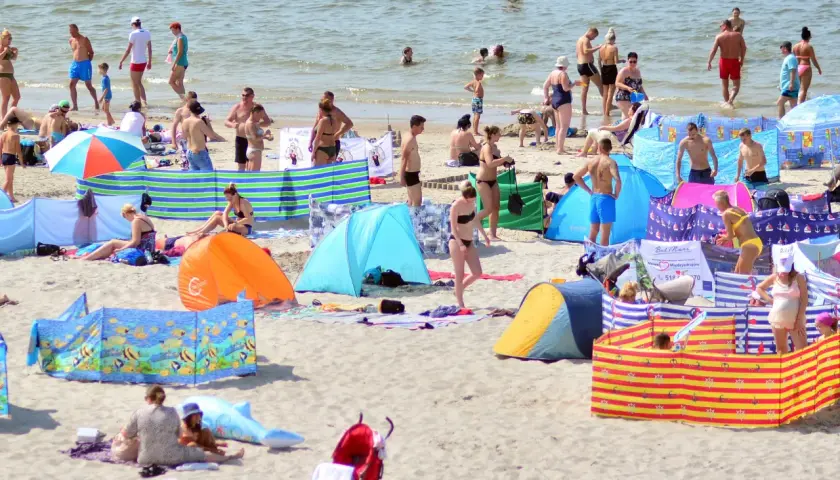Tłum ludzi na plaży, opalający się i kąpiący w morzu, kolorowe parawany i namioty plażowe.