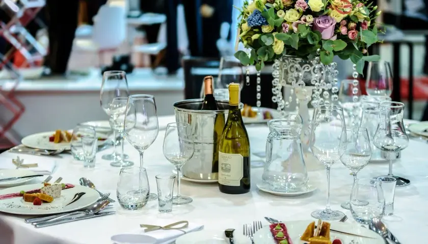 Elegancko nakryty stół w restauracji, gotowy na przyjęcie weselne. Na białym obrusie stoją kieliszki, butelka wina i piękne kwiaty.