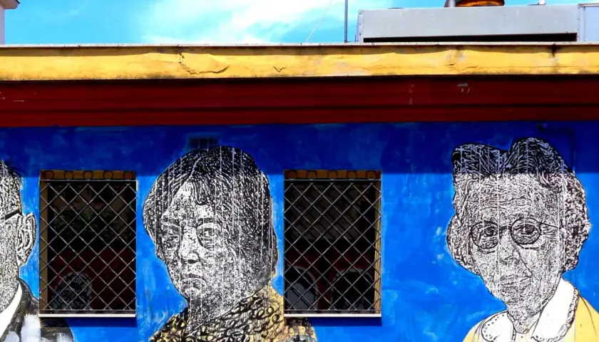Volti stilizzati in bianco e nero su un muro blu acceso, parte di un **murales ostiense**. Le finestre con grate interrompono la composizione.
