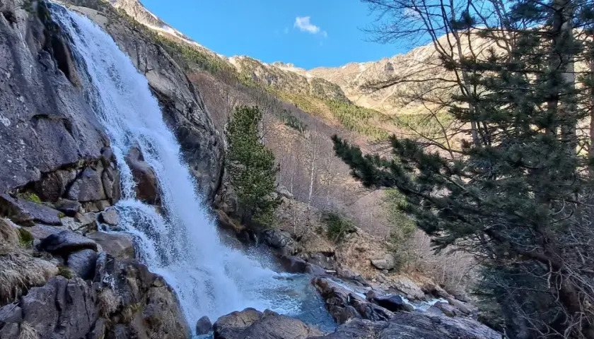 Cascada imponente en un paisaje montañoso, ideal para explorar rutas vielha. El agua cae sobre rocas y vegetación, con árboles y picos nevados al fondo.