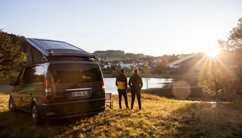 Un camping-car Westfalia avec panneaux solaires, prêt pour l'aventure près de Vulcania. Deux personnes contemplent un lac paisible au coucher du soleil.
