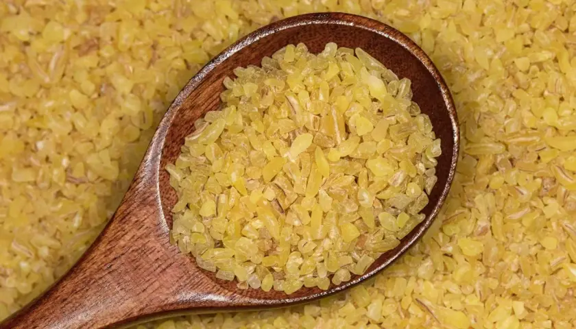 Drewniana łyżka pełna drobnej, żółtej kaszy bulgur na tle rozsypanych ziaren. To zdrowy składnik wielu dań.