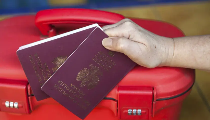 Dłoń trzyma polskie paszporty na czerwonej walizce. Kto może odebrać paszport osoby dorosłej? Tylko ona!