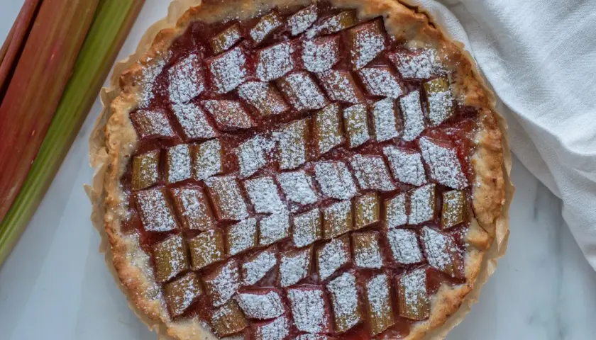 Pyszna tarta z rabarbarem, posypana cukrem pudrem, obok świeże łodygi rabarbaru.