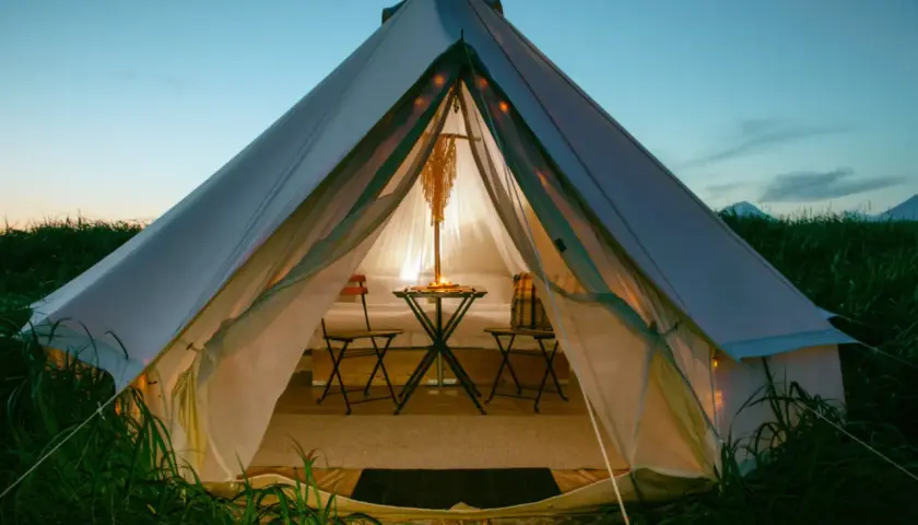 Przytulna glamping jurta oświetlona od środka, z małym stolikiem i krzesłami, otoczona zieloną trawą o zmierzchu.