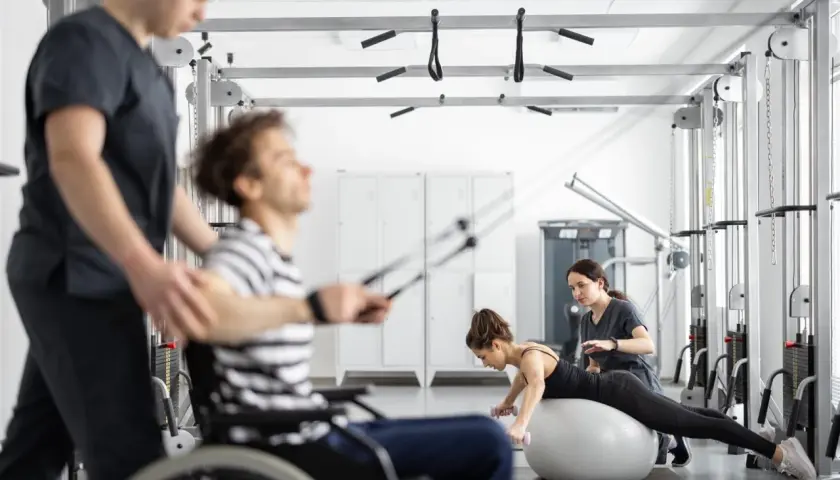 Rehabilitacja metodą Bobath: mężczyzna na wózku ćwiczy z fizjoterapeutą, kobieta na piłce z hantlami pod okiem terapeutki.