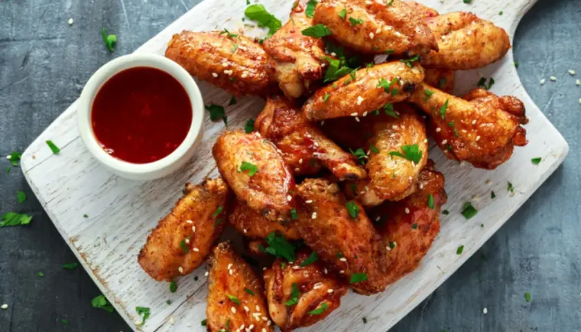 Alitas de pollo al horno jugosas, doradas y espolvoreadas con sésamo y perejil, acompañadas de una salsa roja en un cuenco.