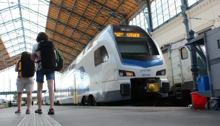 Zwei Reisende mit Rucksäcken warten auf den serbischen Zug nach Szeged unter dem beeindruckenden Dach eines Bahnhofs.