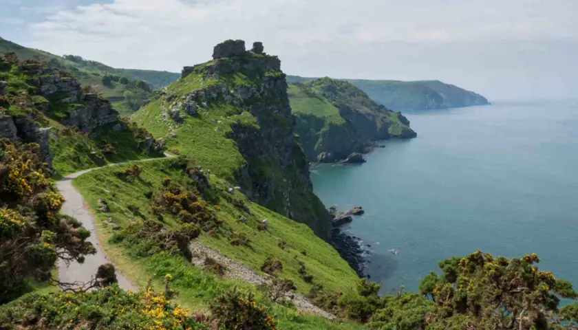A winding path hugs a grassy cliff overlooking the sea. This is the salt path, a scenic route with rugged beauty.