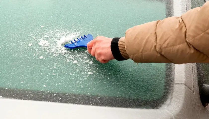 En hand med en blå isskrapa skrapar is från en bilruta för att få isfria bilrutor.