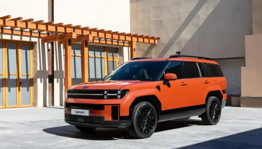 Pomarańczowy, tani SUV Hyundai Santa Fe z czarnymi felgami stoi przed budynkiem z drewnianą pergolą.
