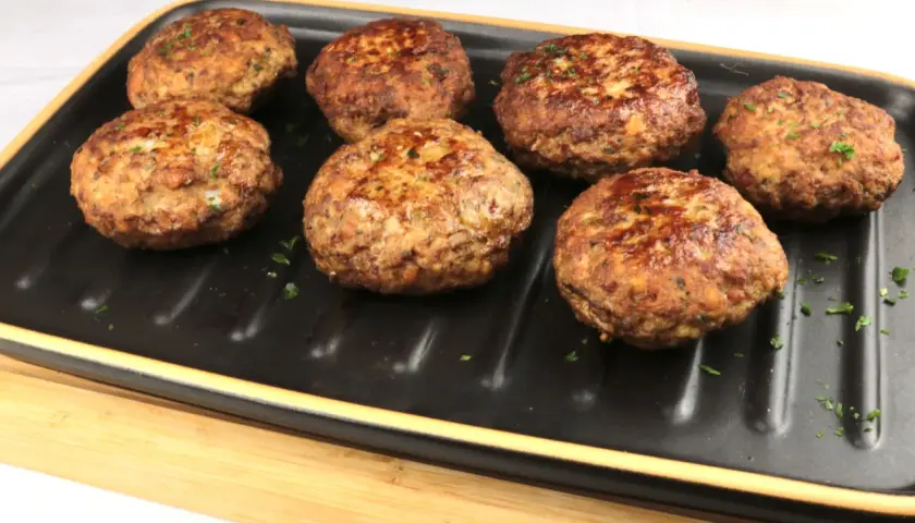 Sieben goldbraune Bouletten, wie nach Omas Rezept, liegen auf einem schwarzen Grillteller, garniert mit Petersilie.