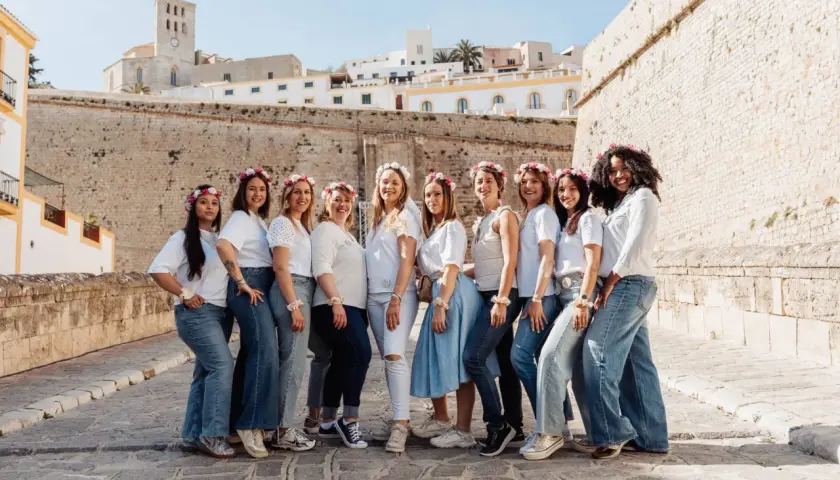 Dix amies célèbrent leur evjf en France, souriantes et parées de couronnes de fleurs, devant un mur de pierre et des bâtiments blancs.