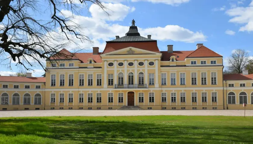 Piękny, żółty pałac z czerwonym dachem, jeden z wielu wspaniałych zamków w Wielkopolsce, otoczony zielonym trawnikiem i drzewami.