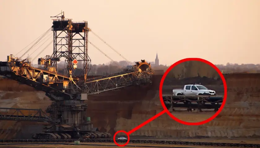 Największa koparka świata w kopalni odkrywkowej. W oddali widać mały biały pickup, który wygląda jak zabawka przy gigantycznej maszynie.