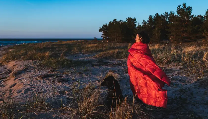 Kobieta w czerwonym śpiworze i pies na plaży. Zastanawiasz się, jaki śpiwór wybrać na nocne wędrówki?