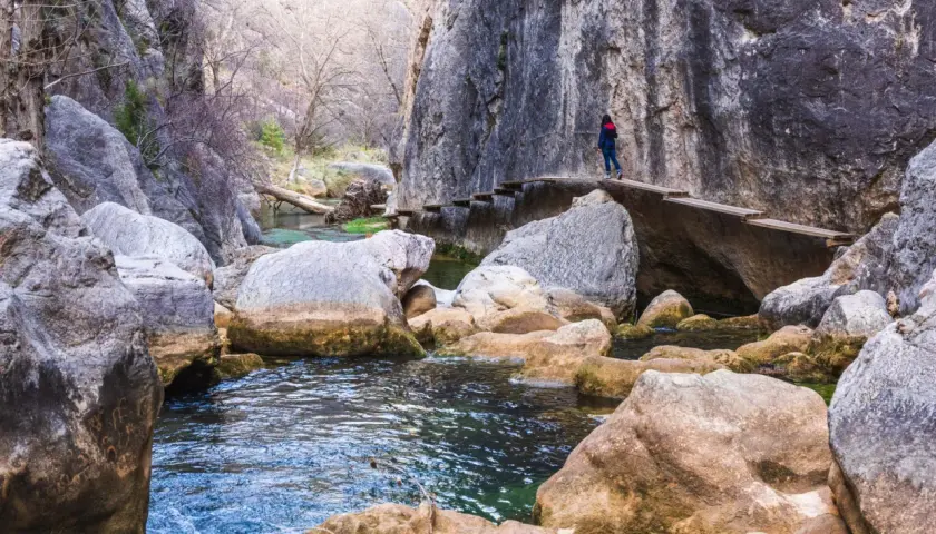 Una persona camina por un puente de madera en la ruta del silencio, rodeada de rocas y un río de aguas cristalinas.
