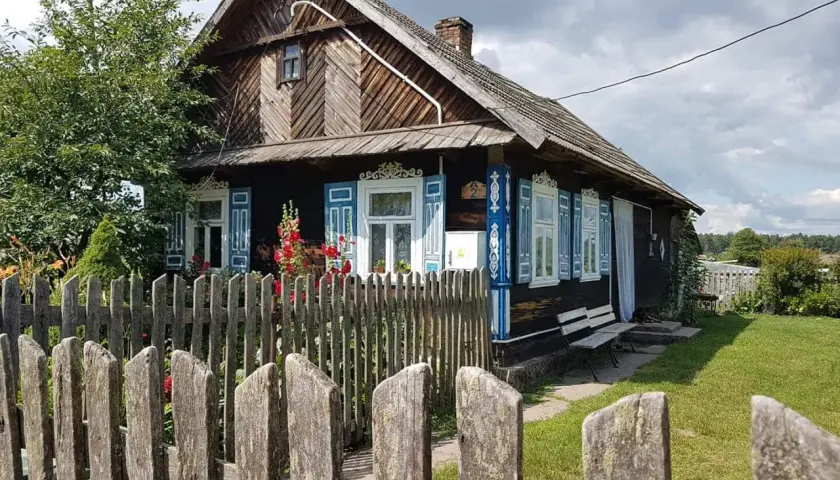 Drewniany dom z niebieskimi, otwartymi okiennicami, ozdobiony ludowymi wzorami. Wokół zielony trawnik i drewniany płot.