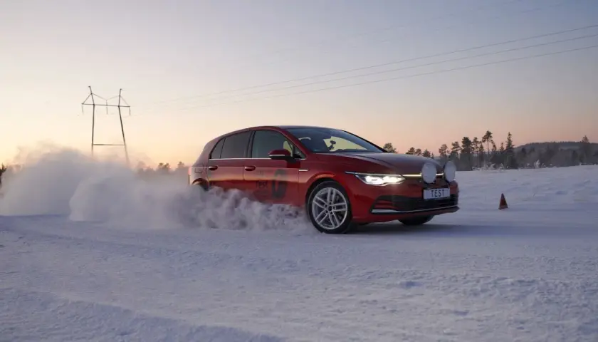 Czerwony Golf podczas kormoran snow test na zaśnieżonej drodze, z chmurą śniegu unoszącą się spod kół.