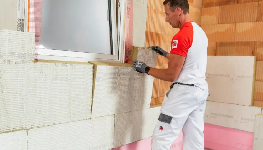Budowlaniec montuje płyty izolacyjne, dbając o ocieplenie domu. Praca przy oknie z użyciem wełny mineralnej.