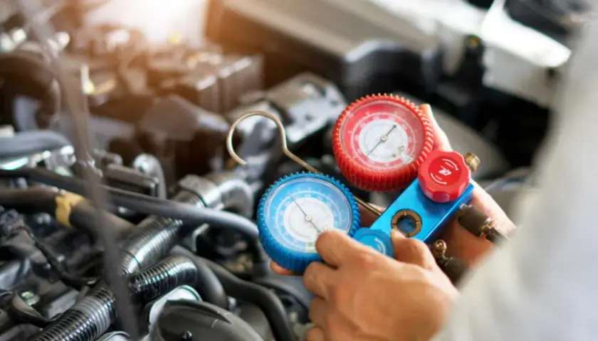 Manometro per ricaricare aria condizionata auto, con due quadranti (rosso e blu) collegati a tubi, in mano a un meccanico davanti al motore.