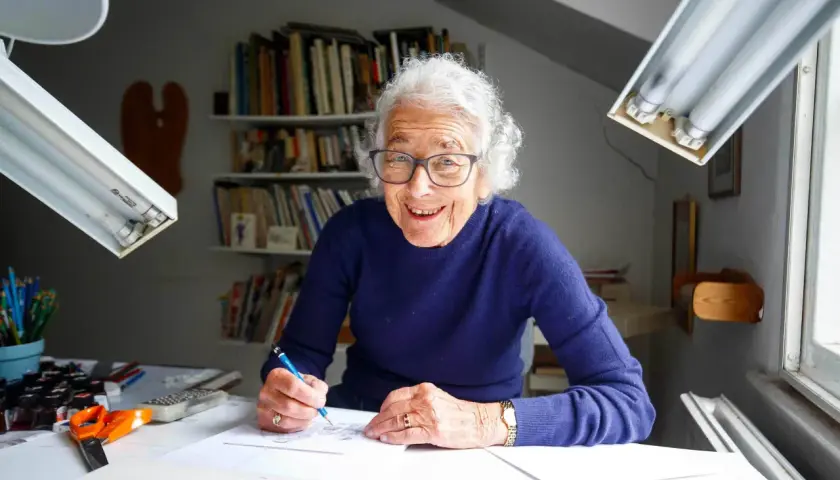 Judith Kerr, lächelnd, zeichnet an ihrem Schreibtisch. Bücherregale im Hintergrund.