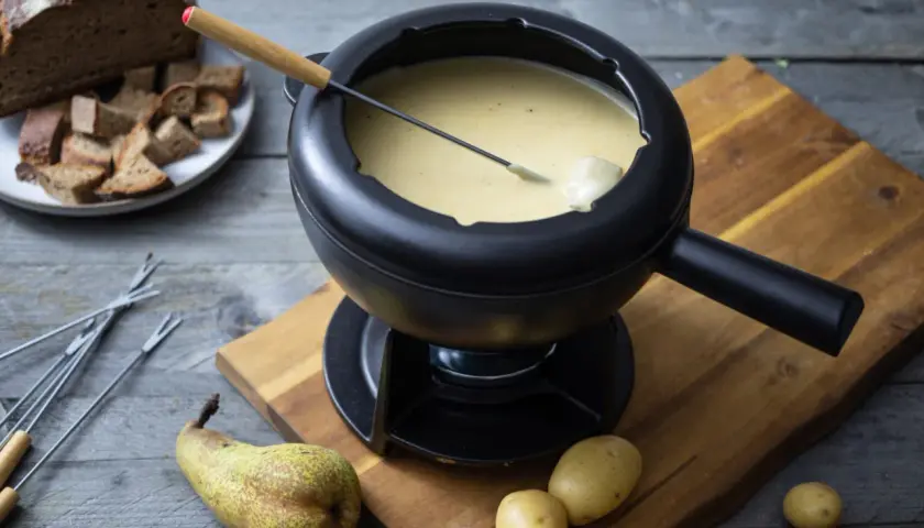 Ein dampfendes Schweizer Fondue in einem schwarzen Caquelon, daneben Brot, Kartoffeln und eine Birne.