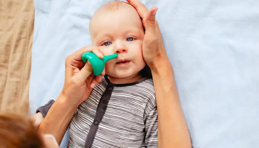 Un bebé con ojos azules recibe un lavado nasal con un aspirador verde. Una mano sujeta su cabeza mientras la otra usa el dispositivo.