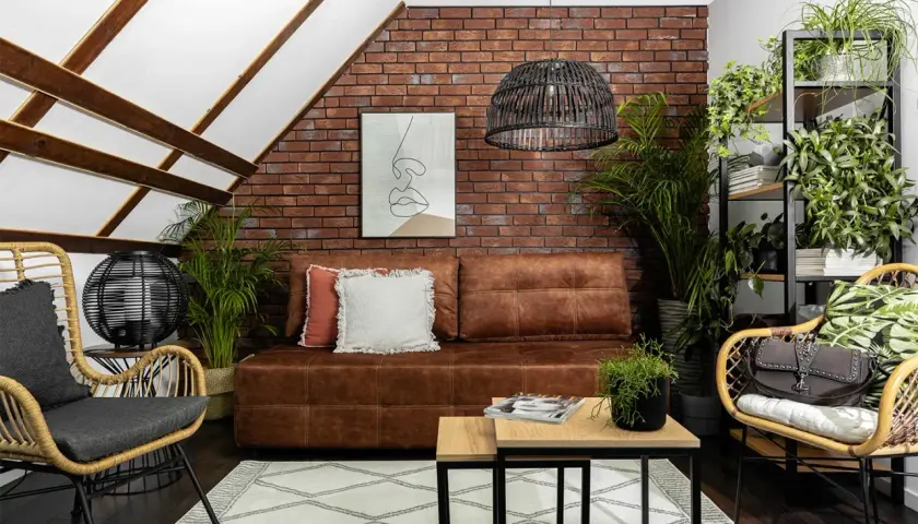 Nowoczesny salon z ceglaną ścianą, na której subtelne oświetlenie ściany z cegły ledami podkreśla jej fakturę. Brązowa kanapa, wiklinowe fotele i mnóstwo zieleni.