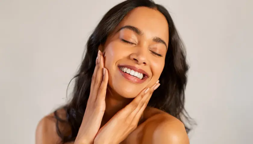 Une femme rayonnante sourit, les yeux fermés, caressant son visage. Ses mains apportent des conseils maquillage pour un teint éclatant.