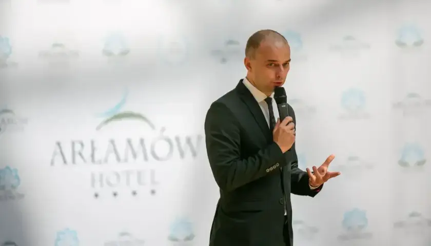 Mężczyzna w garniturze przemawia na tle logo Hotel Arłamów. Właściciel hotelu z pasją opowiada o swoich planach.