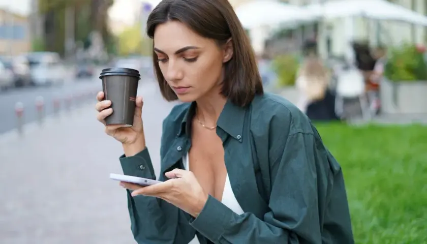 Kobieta z oliwkowym kolorem skóry, trzymająca kawę i telefon, siedzi na ławce w mieście.