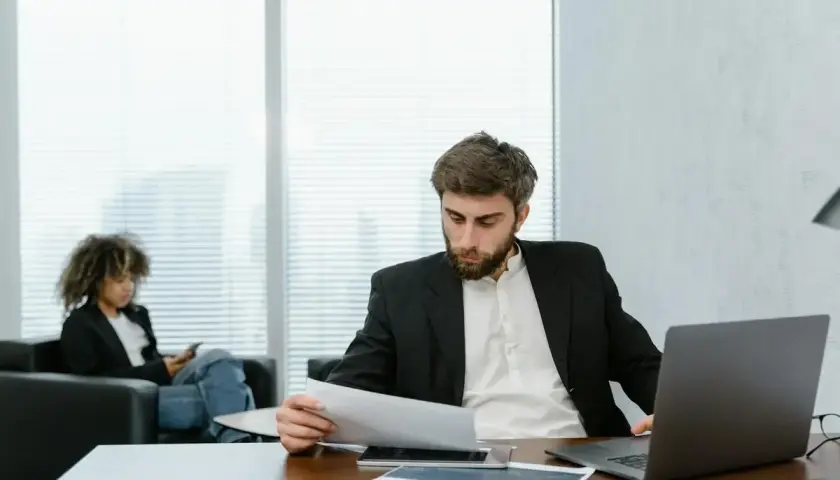 A man in a suit reviews documents, perhaps researching accelerated online MPA programs. A woman in the background is on her phone.