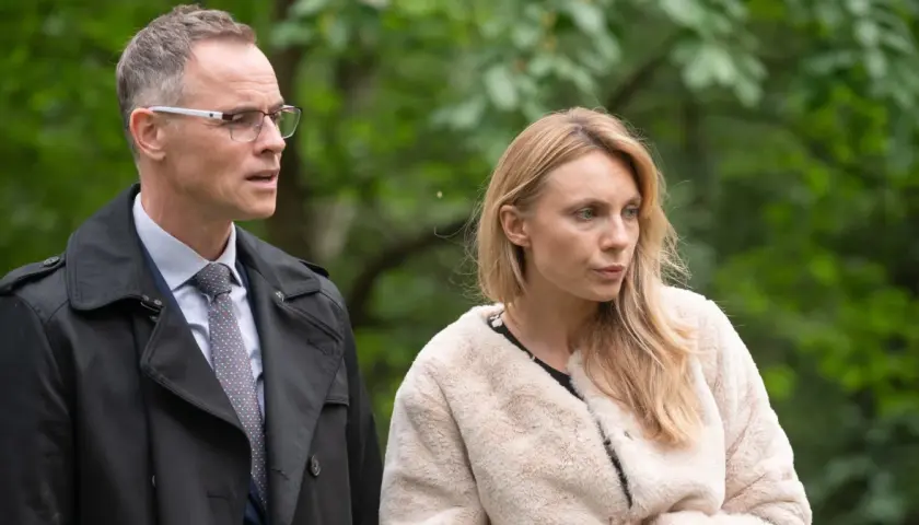 Adam i Ewa obsada: mężczyzna w okularach i płaszczu oraz kobieta w futrzanej kurtce na tle zieleni.