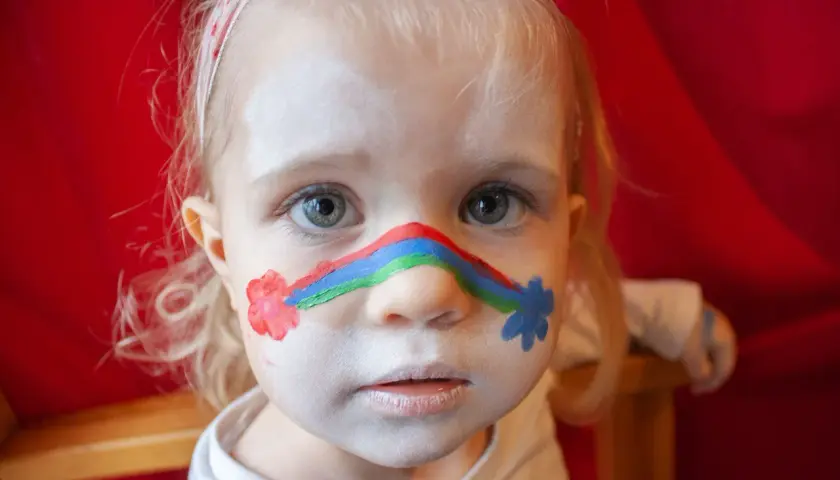 Ein kleines Mädchen mit einem Regenbogen im Gesicht und Blumen auf den Wangen. Kinderschminken einfach gemacht!