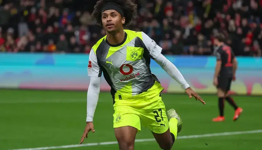 Piłkarz Adeyemi w żółto-szarej koszulce BVB z numerem 27, z afro na głowie, świętuje bramkę na boisku.