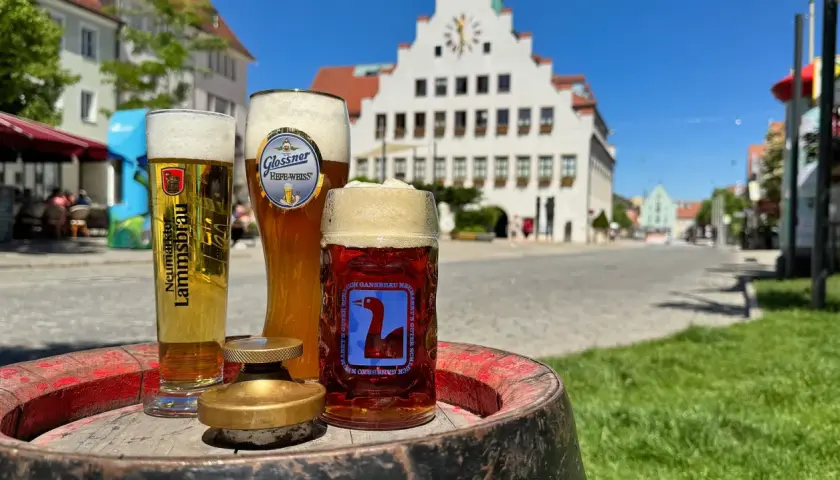 Drei Gläser Bier, darunter ein helles und ein dunkles, stehen auf einem Fass. Im Hintergrund eine malerische Stadtkulisse mit einem historischen Gebäude. Ein perfekter Genuss von italienischem Bier.