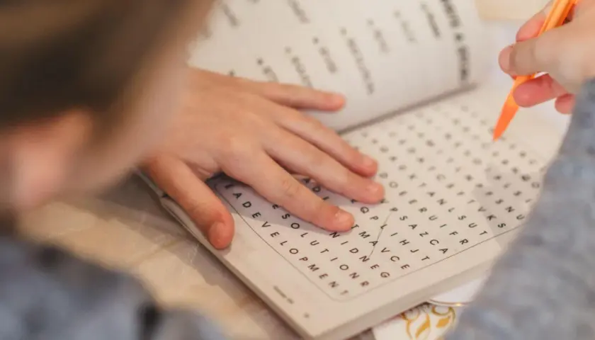 Mano de niño resolviendo una sopa de letras para la memoria con un lápiz naranja.