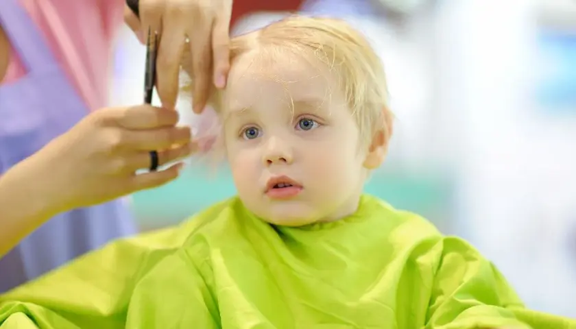 Mały chłopiec z blond włosami otrzymuje swoją pierwszą fryzurę. Włosy są delikatnie cięte, tworząc urocze fryzury dla rocznego chłopca.