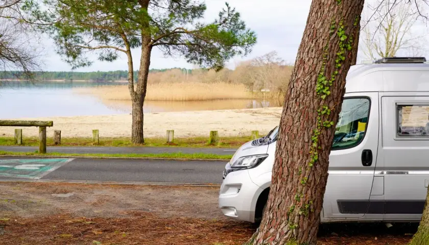 Un fourgon de 6 mètres est garé près d'un lac, à l'ombre d'un grand arbre. Le paysage est paisible, avec une plage de sable et des roseaux au loin.