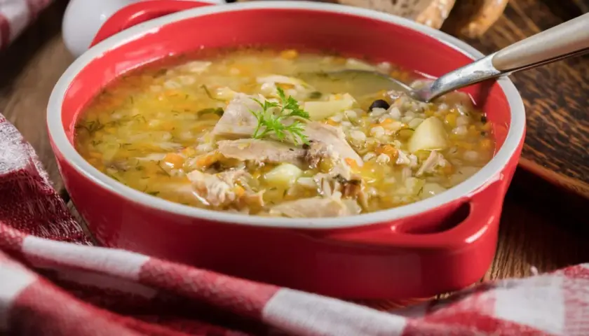 Gorąca zupa krupnik przepis babci w czerwonej misce, z kawałkami mięsa, ziemniaków i kaszy pęczak. Obok kromki chleba i czerwono-biała serwetka.