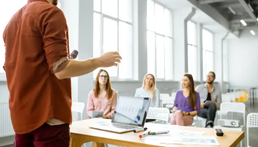 Mężczyzna z mikrofonem prezentuje swój elevator pitch grupie słuchaczy. Na stole laptop z wykresami.