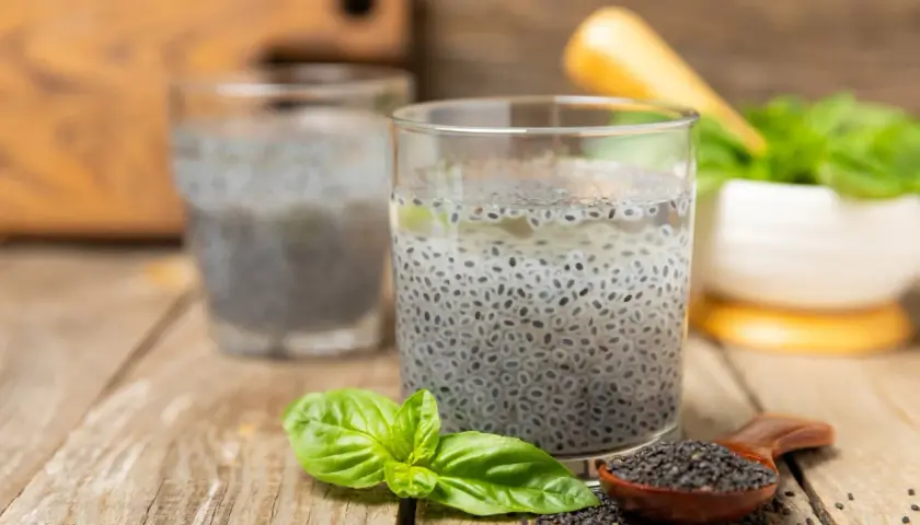 Due bicchieri con semi di basilico in acqua, un cucchiaino di semi e foglie fresche su un tavolo di legno.